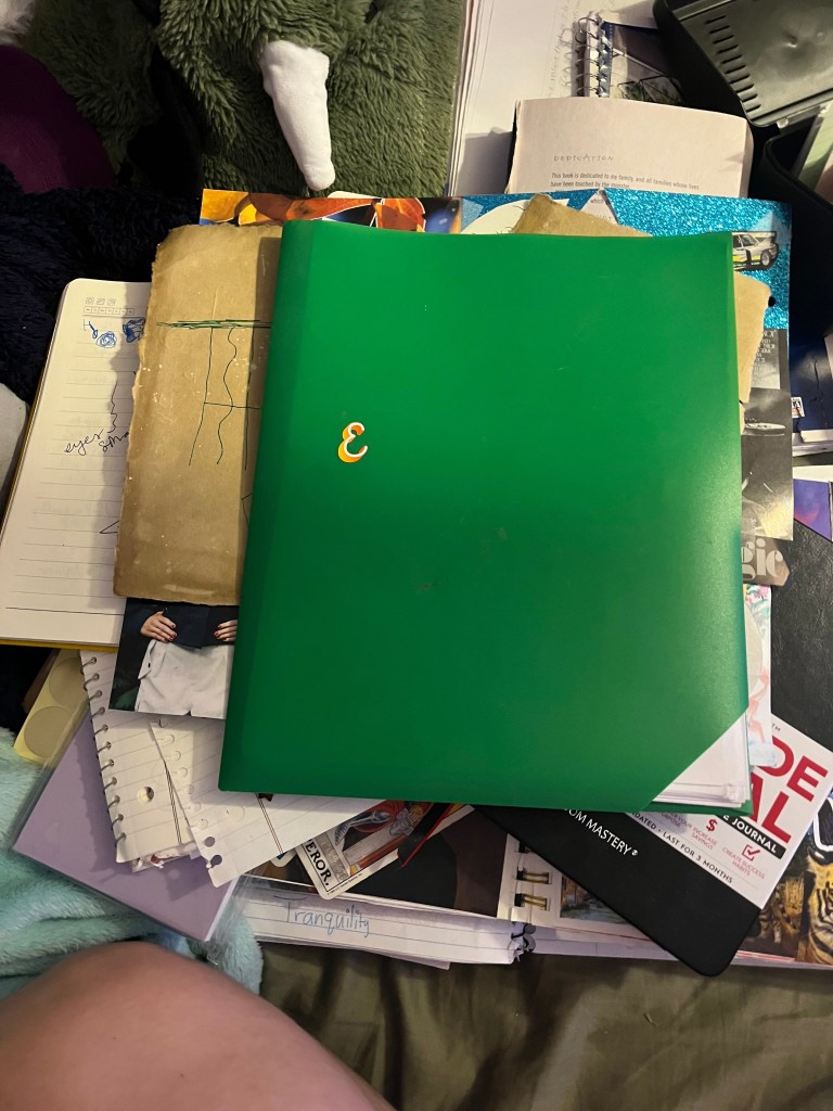 A cluttered workspace featuring a stack of various notebooks, papers, and a green folder labeled with the number '3'. A cozy blanket is visible as part of the scene.