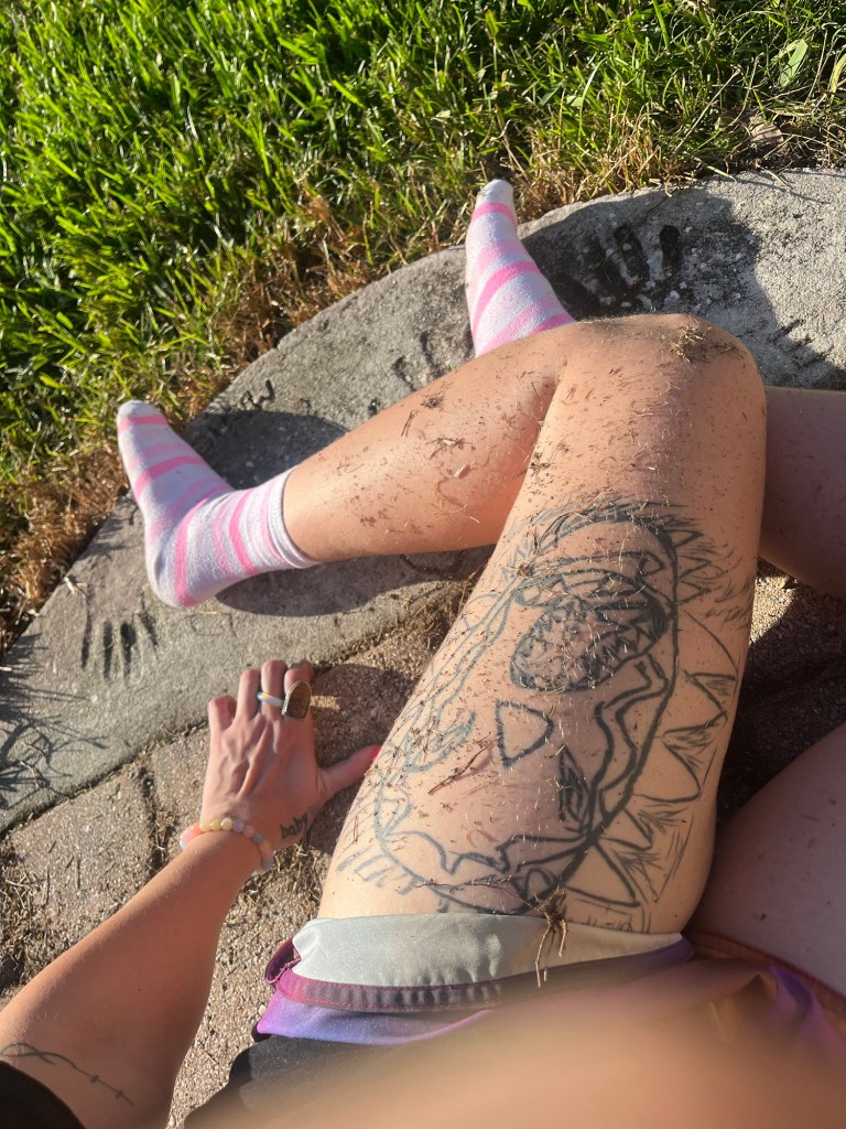 A close-up of a person's legs with visible tattoos, sitting on a stone surface. The legs are covered in dirt and grass, and the person is wearing pink and white striped socks. A hand is placed on the thigh, and a handprint is visible on the stone.