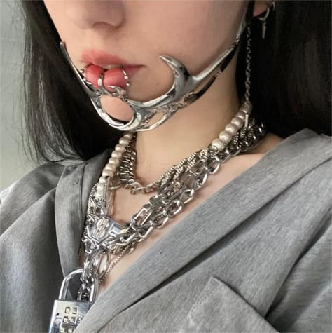 A close-up portrait of a person with dark hair wearing a gray hoodie. They have a unique silver facial accessory that covers part of their mouth and chin, along with multiple silver and pearl necklaces layered around their neck.