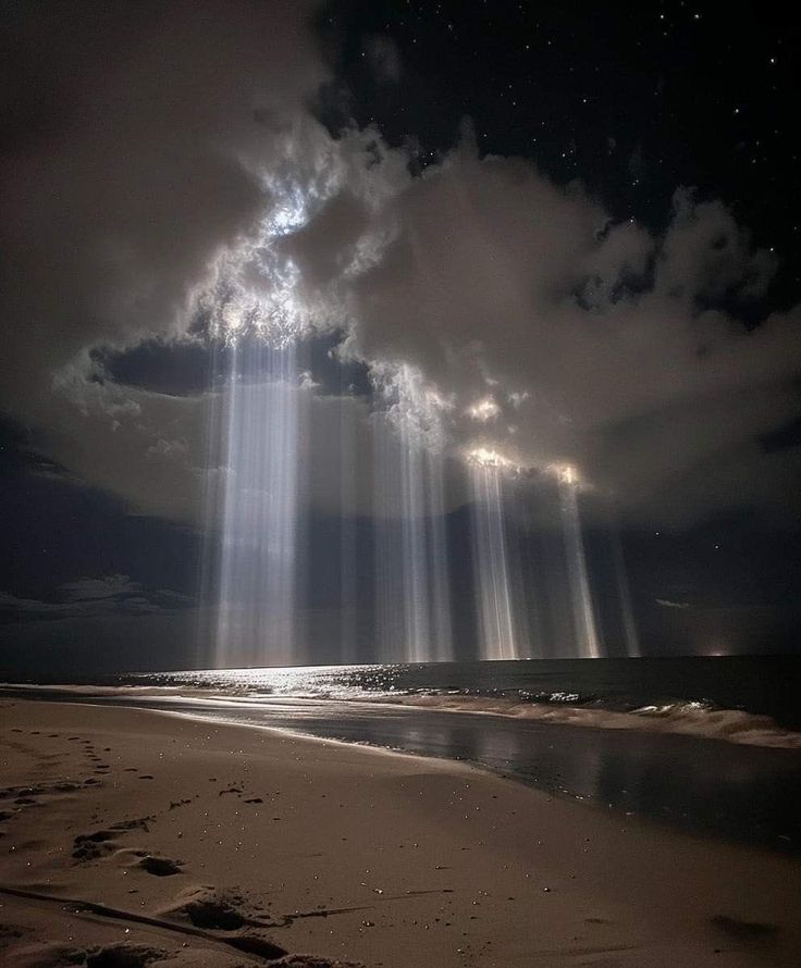 A night sky with beams of light shining through clouds above a calm ocean, with a sandy beach in the foreground.
