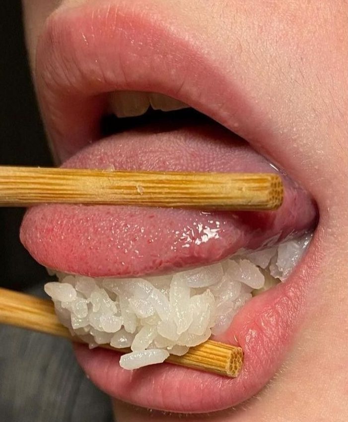 Close-up of a mouth holding sushi rice between chopsticks.