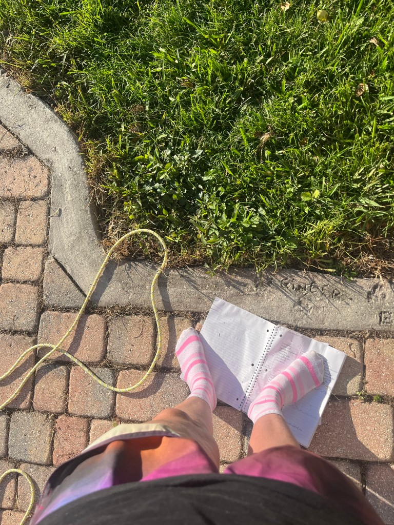A person's feet in pink striped socks standing on a sheet of paper on a patterned pavement next to green grass and a yellow rope.