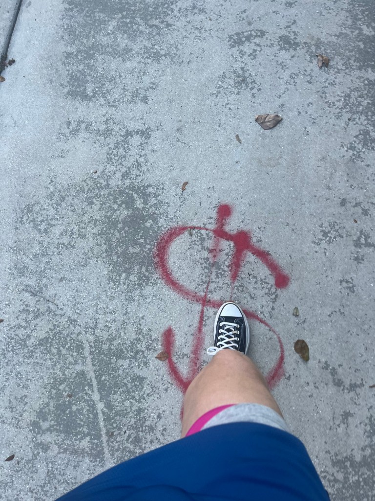 A foot in a black sneaker stepping over a red spray-painted dollar symbol on a concrete surface, with some fallen leaves scattered around.