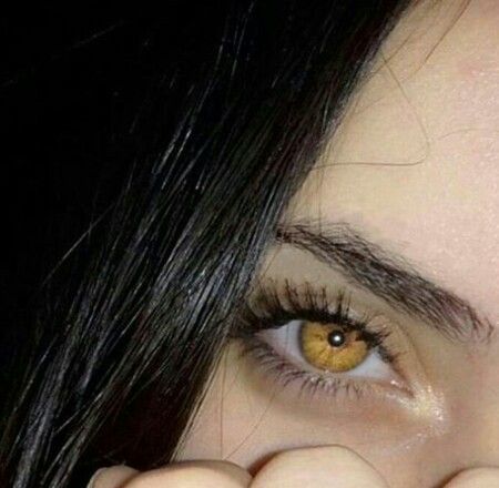 Close-up of a person's eye with golden-brown iris and long eyelashes, partially obscured by a hand.
