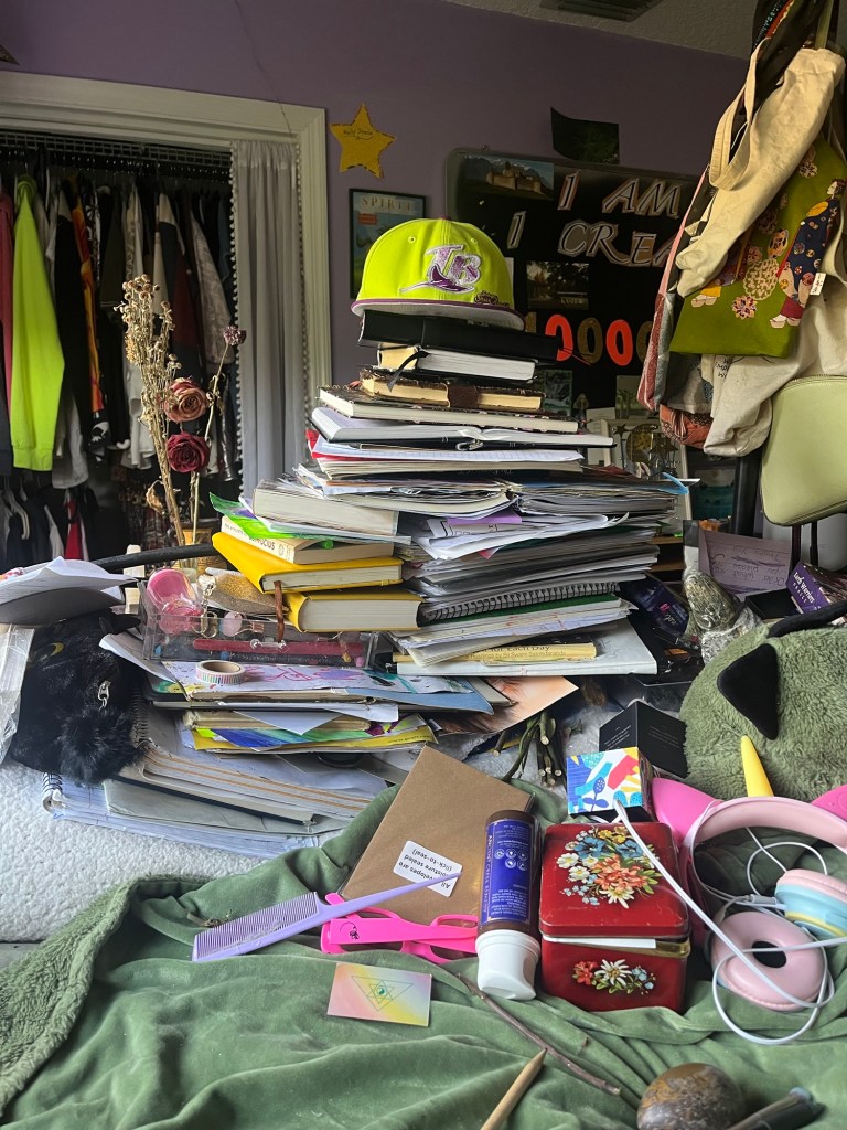 A cluttered workspace featuring a tall stack of books and notebooks, a bright yellow cap on top, and various items like a comb, a decorated tin, and electronic headphones scattered around.