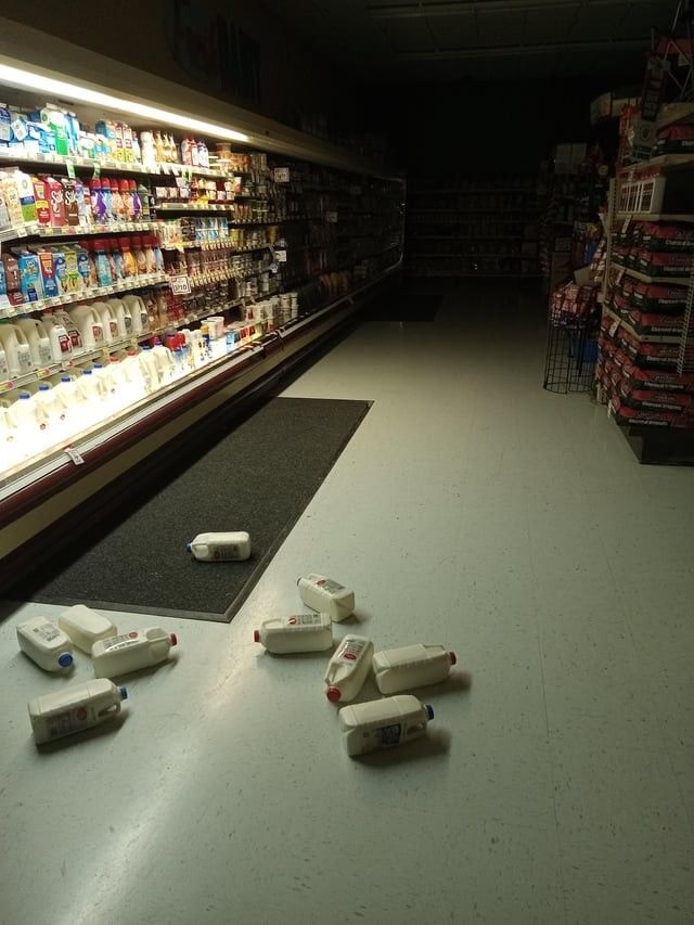 An aisle in a grocery store featuring a variety of dairy products on display, with several milk jugs toppled and scattered across the floor.