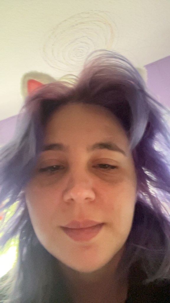 Close-up selfie of a person with purple hair and cat-ear headband, taken indoors with a soft background.