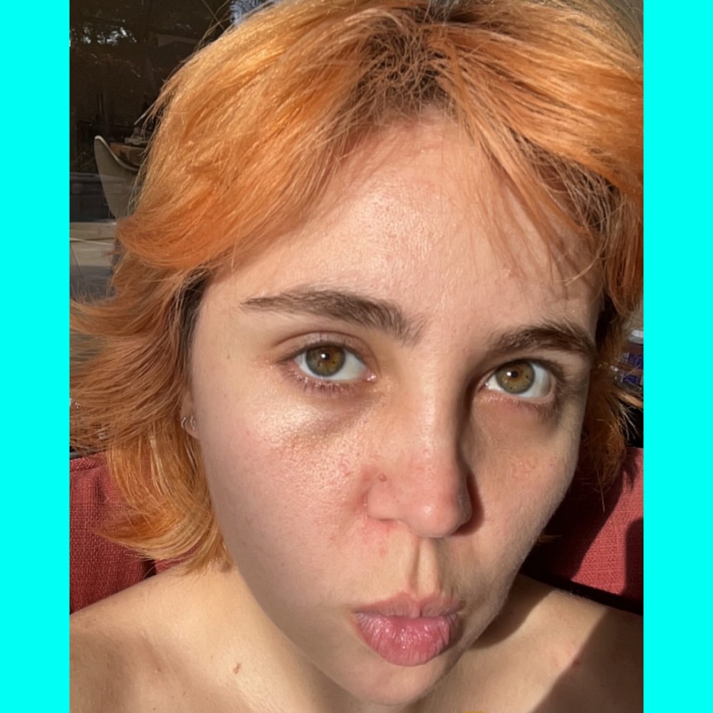 A close-up portrait of a person with short, orange hair, looking directly at the camera with a thoughtful expression, against a bright turquoise background.